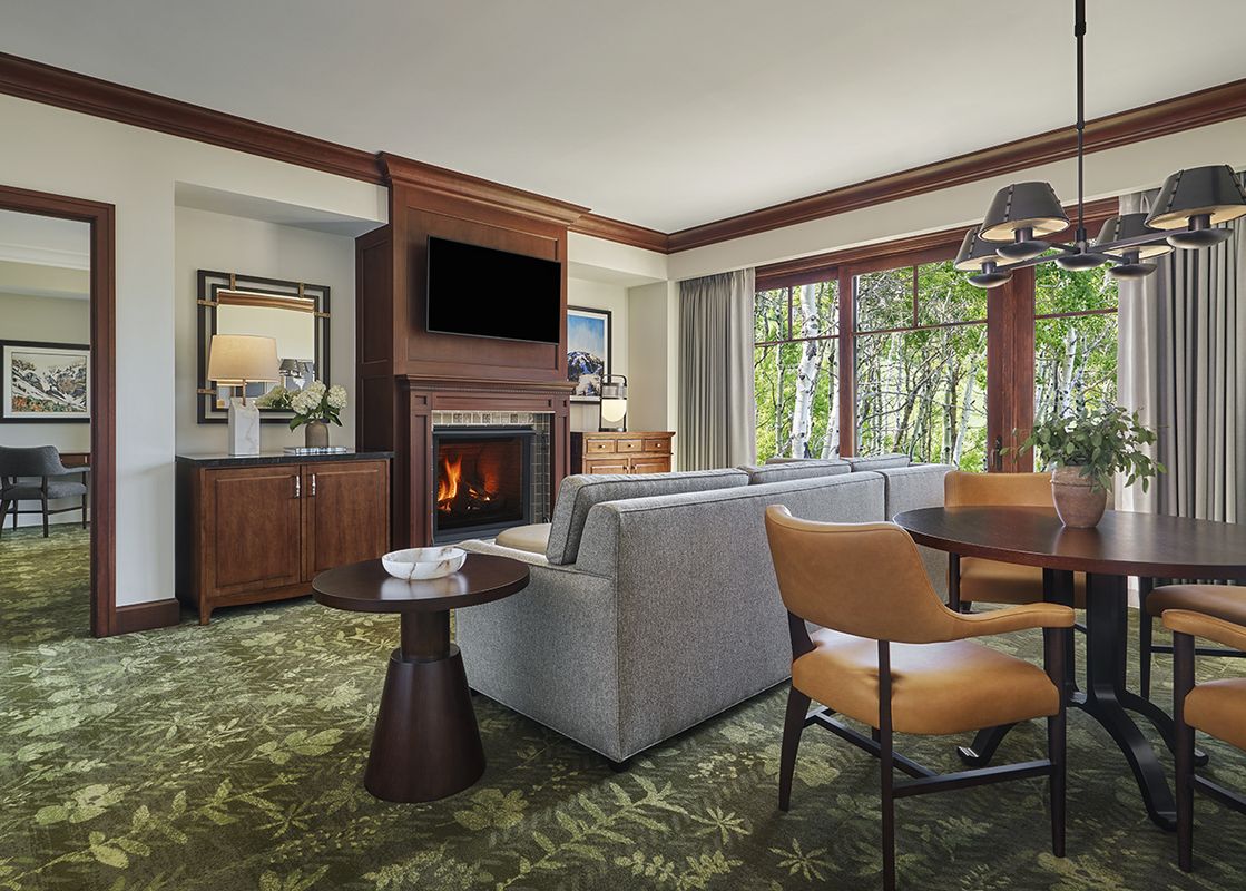 A spacious and elegantly furnished living area inside the Montage Deer Valley Accessible Resort View King Suite, with a fireplace, comfortable seating, and a dining table, featuring large windows that offer a view of green trees outside.