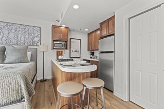 A cozy studio apartment with a bed on the left and a kitchenette on the right, featuring modern appliances and a breakfast bar.