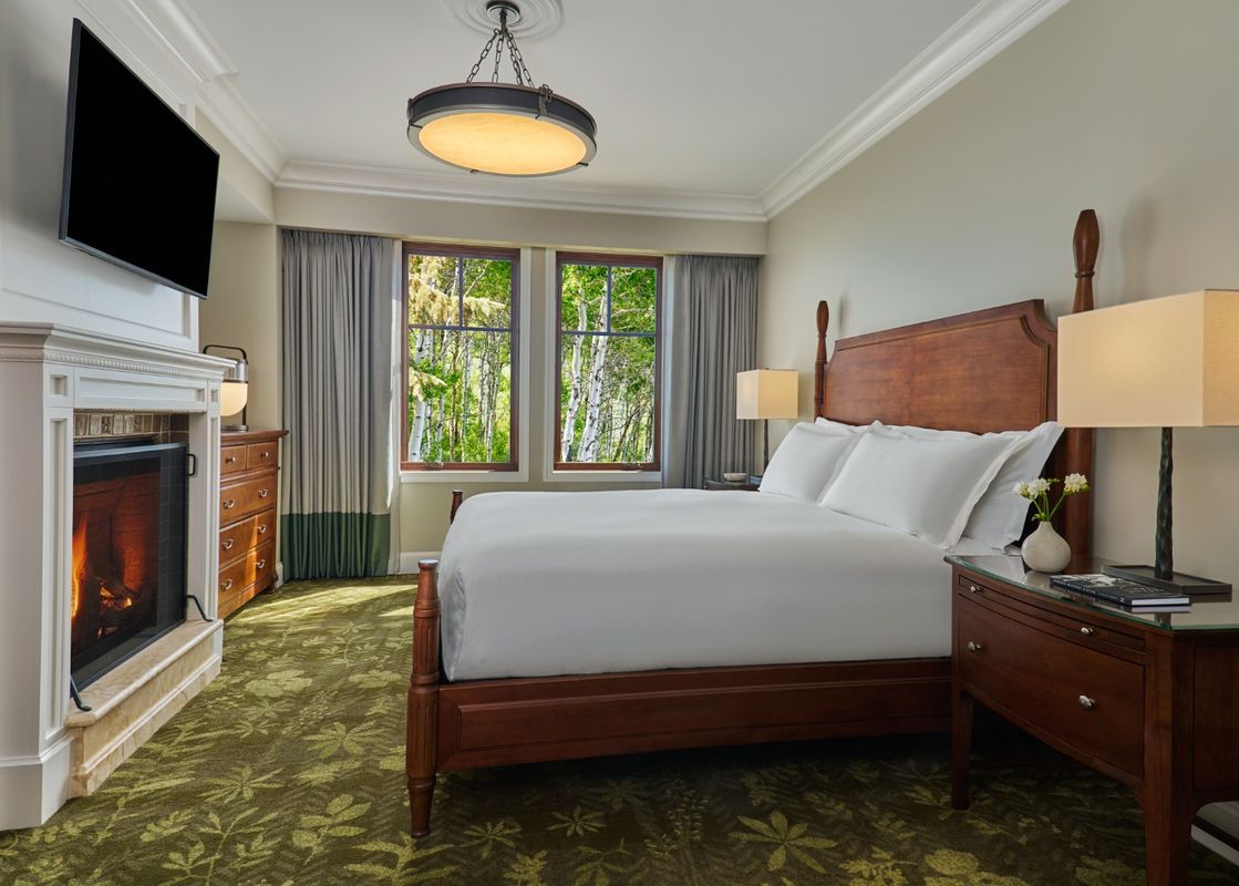 A spacious bedroom with a large wooden bed, a lit fireplace, and two windows offering a view of lush green trees.