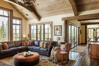 A luxurious living room with a large leather sectional, an ottoman, and a fireplace, featuring large windows with mountain views and a high wooden ceiling.