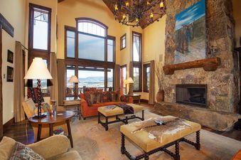The living area at 492 Golf Course Circle boasts a grand stone fireplace, a large chandelier, and expansive windows.