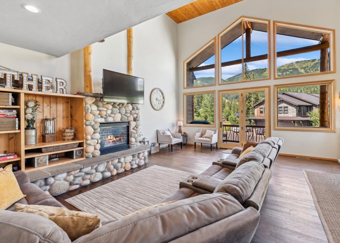 A spacious living room with a large stone fireplace, a flat-screen TV mounted above, a comfortable sectional sofa, and expansive windows offering stunning mountain views.