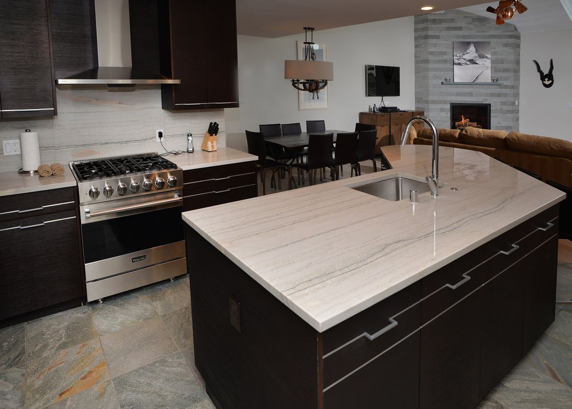A modern kitchen with a large island, stainless steel appliances, dark wood cabinetry, and a view of a dining area and living room with a fireplace.