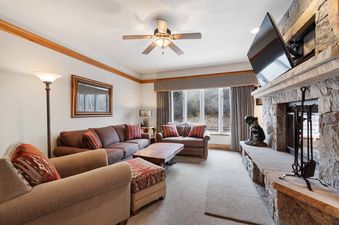 The lounge at The Charter D2705 features comfortable brown sofas, a rustic wooden coffee table, a stone fireplace with a mounted TV, and large windows with checkered curtains, creating a warm and inviting atmosphere.