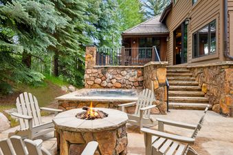 A welcoming outdoor entertainment area at Winter Watch Estate, featuring a lighted fire pit, hot tub, and stone staircase leading to a balcony, all set against a backdrop of dense pine trees.