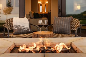Two wine glasses sit on a tree stump table between two chairs on an outdoor patio with a fire pit in the foreground and an open suite in the background.