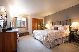 A spacious bedroom at Chateau Chamonix CX213, featuring a large bed with patterned bedding, bedside lamps, a dresser with an oval mirror, and a window with a view of trees.