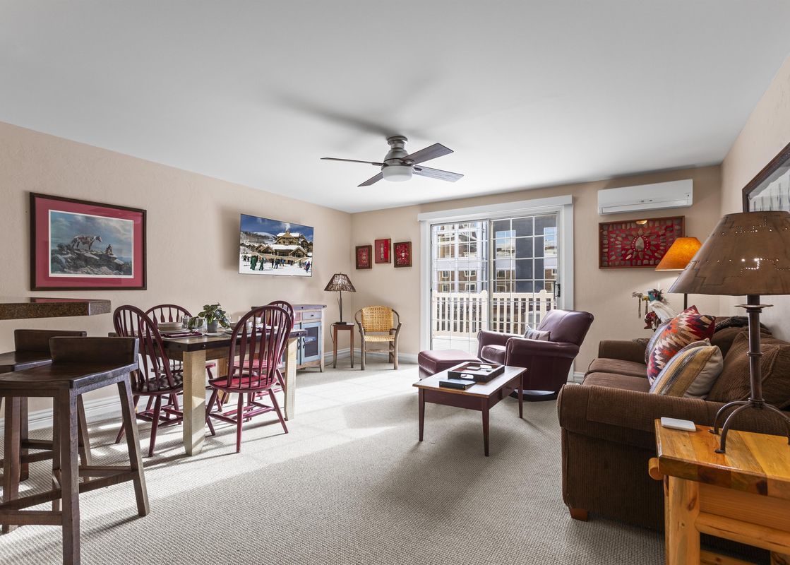 A bright living and dining area with light-colored walls, carpeted floors, and a mix of wooden and upholstered furniture.