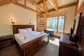 A spacious bedroom featuring a king-sized bed with white bedding, wooden furniture including a dresser and nightstand, and large windows offering a view of the outdoors.