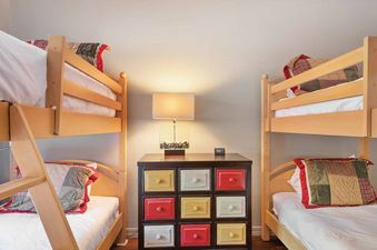 A cozy bedroom features two wooden bunk beds with white linens and colorful patterned pillows, separated by a dark wood dresser with nine multicolored drawers and a modern lamp.