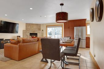 An open-concept living room with a brown leather sofa, a stone fireplace, a dining table set for guests, and a modern kitchen.