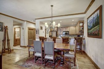 A spacious dining room features a large wooden table with eight upholstered chairs, a vintage chandelier, and hardwood floors with a decorative rug, all opening into a modern kitchen area.