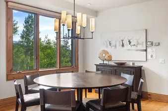 A round wooden dining table surrounded by eight upholstered chairs sits in a bright dining room with a large window overlooking a lush green mountain forest.