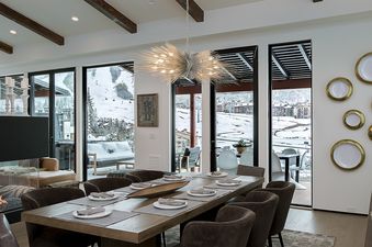 A modern dining room with a large wooden table set for eight, a sputnik chandelier, and floor-to-ceiling windows offering a snowy mountain view.