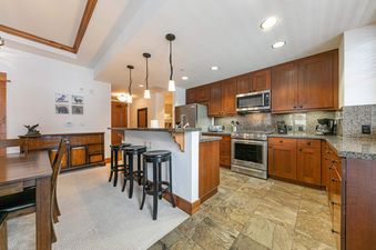 Big Horn Lodge 202 kitchen has stainless steel appliances, granite countertops, and a breakfast bar with three stools. The kitchen is connected to the dining area where there is a large wooden table and chairs.