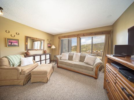 The living area at Snowblaze E310 features a comfortable sofa and armchair, a wooden dresser with a television, and a large window offering a view of the mountains.