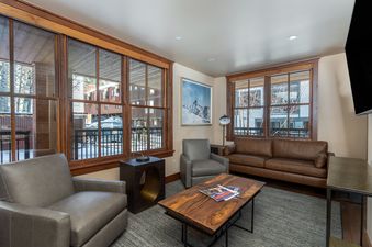 A cozy living room in Ballard Haus with two gray armchairs, a brown leather sofa, a wooden coffee table, and large windows offering a snowy outdoor view.