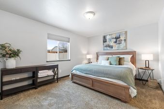 A serene bedroom at Destiny Cove features a wooden bed with plush bedding, an abstract painting, a dark wooden console table with flowers, and a large window providing natural light.