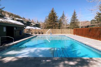 An outdoor swimming pool at Lakeside 1629 is shown with clear blue water, surrounded by paved decking and a gabion wall, under a bright blue sky.