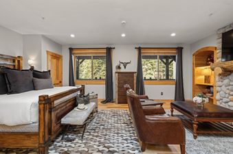 A cozy bedroom with a wooden bed, patterned rug, leather armchairs, and windows overlooking a forest.