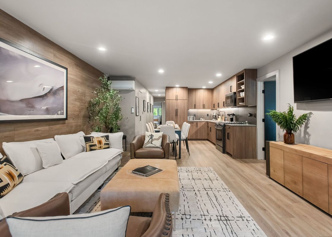 A modern and spacious open-concept living area in Timber Ridge 1B features a comfortable white sectional sofa, a brown leather armchair, a rectangular ottoman, and a long wooden console table.