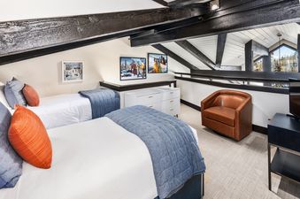 The loft bedroom in Crestwood 3212 Premier 3 Bedroom Plus Loft features two twin beds, each adorned with white linens and blue quilted blankets, complemented by orange and grey throw pillows, set against a backdrop of angled dark wooden beams and a white