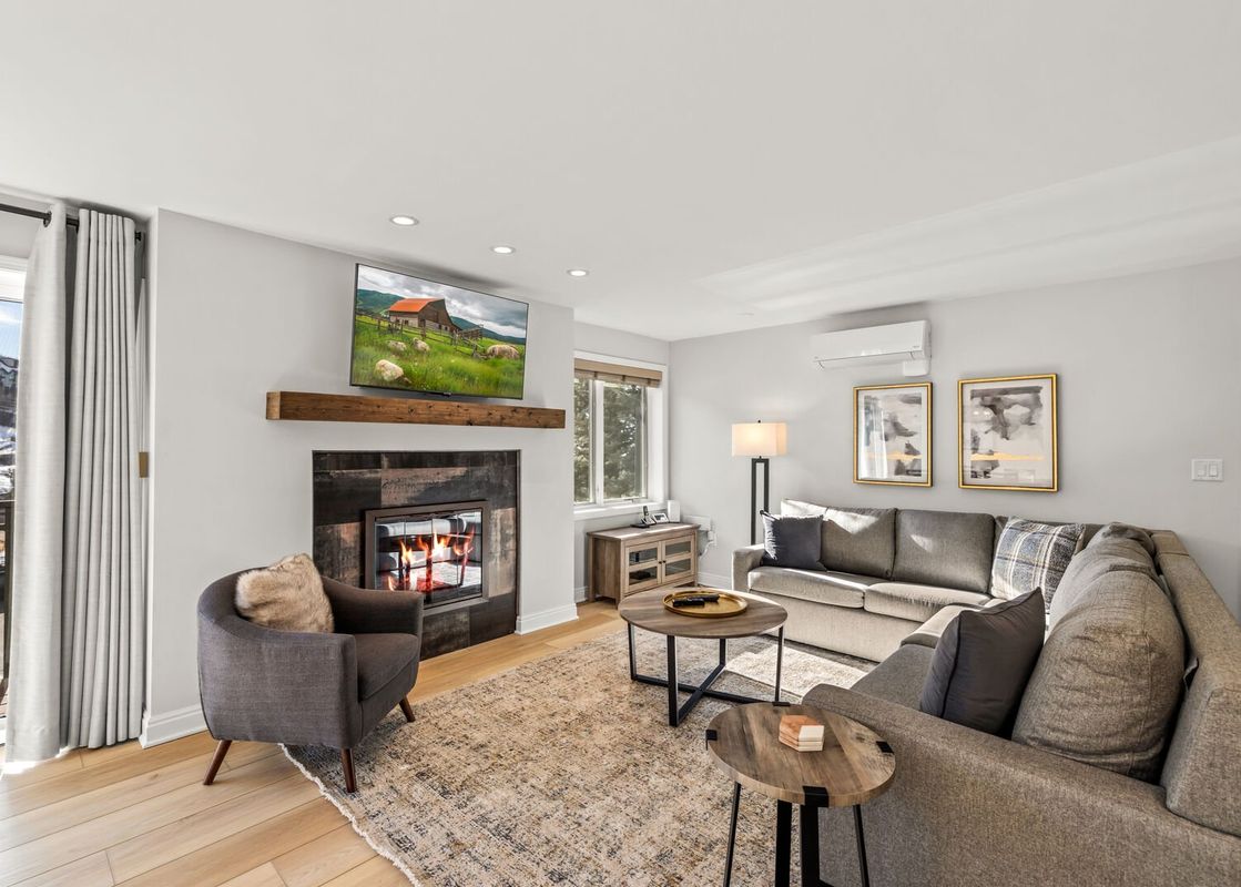 A cozy living room in Bear Claw 2 Valley View 207 featuring a comfortable L-shaped sofa, an armchair, two coffee tables on a rug, a fireplace with a TV above it, and bright natural light from a window and glass door.