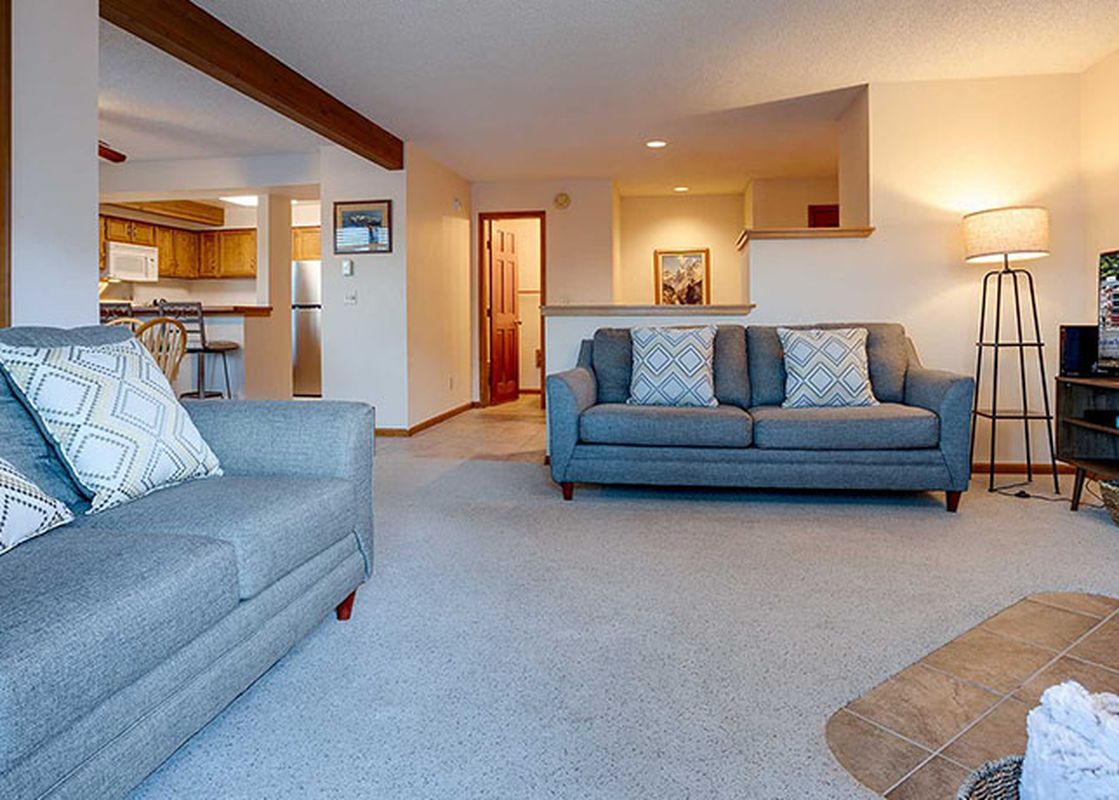 The comfortable living area at Ranch at Steamboat RA411 features two gray sofas with patterned pillows, a large TV, and a floor lamp, with a view of the kitchen in the background.