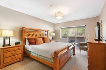 A cozy bedroom at St James Place 235 with a large wooden bed, two bedside tables with lamps, a dresser with a TV, and a sliding glass door leading to a balcony.