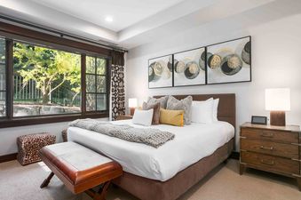 A serene bedroom in Flagstaff 208 features a king-size bed with white linens, various pillows, a knit throw, wooden nightstands with lamps, abstract wall art, and a large window overlooking a lush exterior.