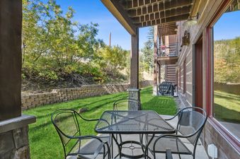 An outdoor patio at Pines Unit 120 with a black metal dining set, a green lawn, and a retaining wall, surrounded by trees under a clear blue sky.
