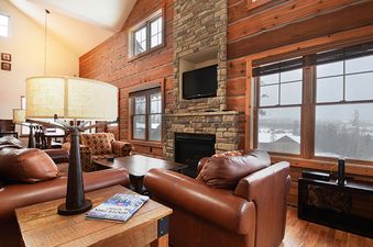 The living area of Rendezvous 25 Discovery Lane features comfortable leather sofas, a stone fireplace, and large windows offering a snowy outdoor view.