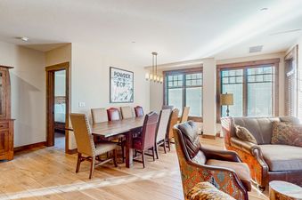 A modern living room with a large wooden dining table and chairs, leather furniture, and a view of the mountains.