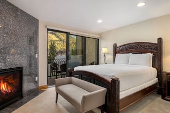 A spacious king bedroom features a dark wood bed with intricate carvings, a stone fireplace, and a private balcony overlooking mountains.