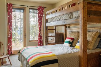 A rustic bedroom in Docs Holiday features wooden bunk beds with colorful bedding and striped duvets, and red star-patterned curtains frame glass doors leading to a pine forest view.
