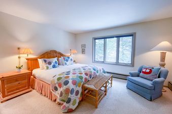 A cozy bedroom with a colorful quilt on the bed, a woven bench, a blue armchair, and a window with a forest view.