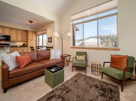 The inviting living area of Trailhead Lodge 731, featuring a comfortable brown leather sofa, green armchairs, and large windows offering a snowy mountain view.