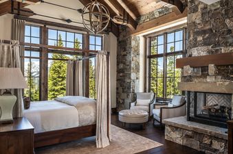 A king-size bed with a canopy is in the center of a luxuriously furnished bedroom in 7 Renegade Rd Moonlight Basin Lodging.  The bed has a white duvet and beige patterned throw pillows.  Two large windows with views of pine trees and a snow-capped mountai