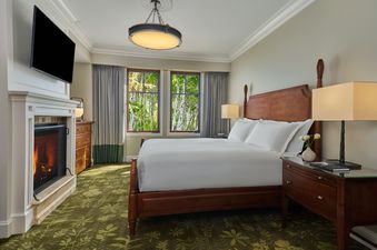 A spacious bedroom with a large wooden bed, a lit fireplace, and two windows offering a view of lush green trees.
