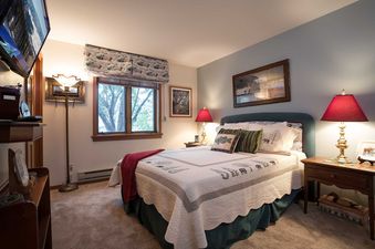 A cozy bedroom with a patterned quilt, wooden furniture, red-shaded lamps, and a window overlooking greenery, featuring a TV on the left.