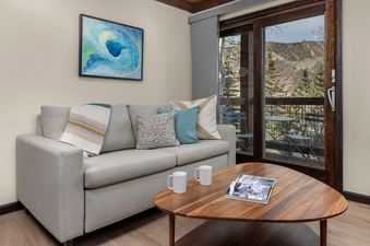 A cozy living room at Willows C3 features a light grey sofa with decorative pillows, a wooden coffee table, and a vibrant abstract painting on the wall, with glass doors opening to a balcony with mountain views.