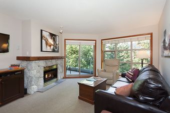 A cozy living room at Aspens on Blackcomb Unit 415 features a stone fireplace, comfortable leather sofa, and large windows offering views of lush greenery.