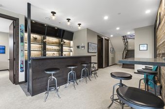 A modern entertainment bar with a dark countertop and stools, backed by rustic wooden panels, illuminated shelves with glassware, and flat-screen TVs.