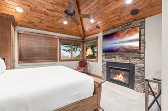 A cozy bedroom with a wooden bed, stone fireplace, flat-screen TV, and large windows with wooden blinds and a view of trees.