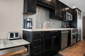 The modern kitchen features dark cabinetry, light countertops, a metallic tile backsplash, and stainless steel appliances, including a coffee maker, toaster, microwave, dishwasher, and range, all set against a warm hardwood floor.