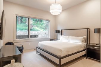 The bedroom at Moonlight Basin 80B Lodgeside features a large bed with white linens, a view of the forest from the window, and a black dresser with a woven basket beside it.