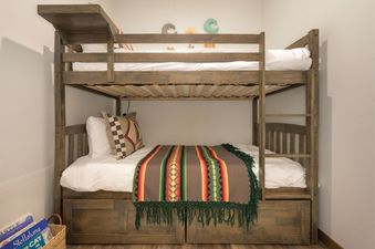 A sturdy wooden bunk bed with white bedding, a colorful blanket, and a decorative pillow.