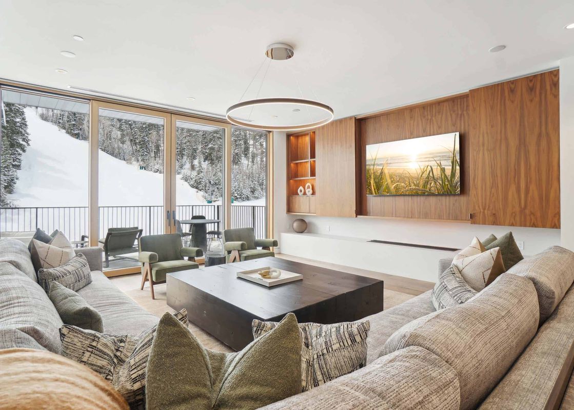 A modern living room with a large sectional sofa, two olive green armchairs, and a dark wooden coffee table, featuring floor-to-ceiling windows overlooking a snow-covered mountain slope with pine trees.