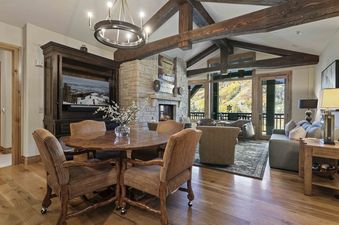 A rustic-chic living and dining area with a stone fireplace, exposed wooden beams, and expansive windows offering mountain views.