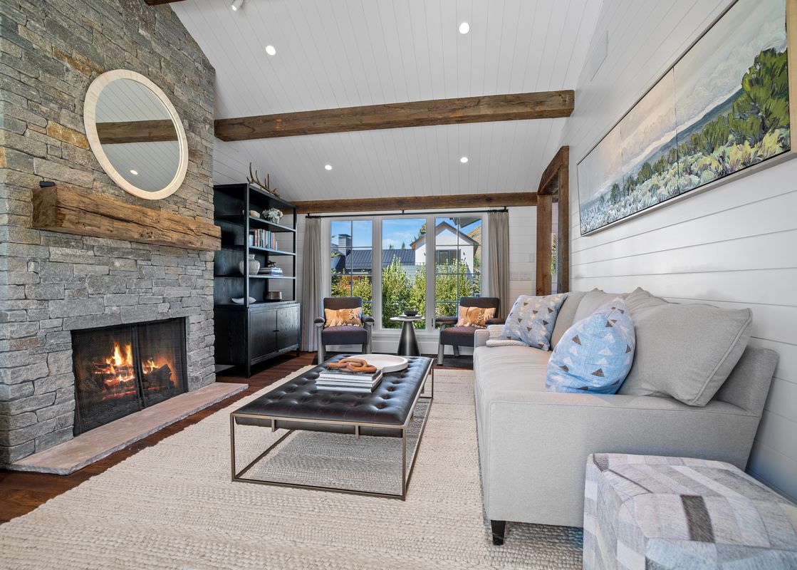 A cozy living room featuring a stone fireplace with a fire, a light gray sofa with throw pillows, a black leather ottoman, and built-in shelving, under a white shiplap ceiling with wooden beams.